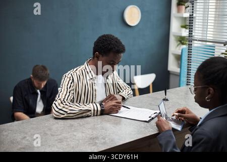 Schwarzer junger Mann, der Papiere am Bankschalter ausfüllt, während schwarze Angestellte mit Personalausweis ihm helfen, männlicher Kaukasier sitzt im Hintergrund und wartet Stockfoto