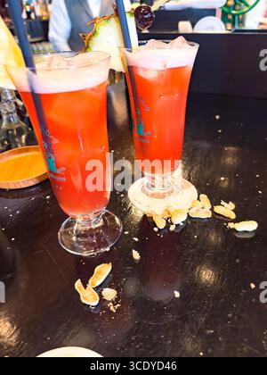 Zwei leuchtend rote Singapore Sling Cocktails, garniert mit Ananas und Kirschen, sitzen in der Long Bar des Raffles Hotel mit verstreuter Erdnusstradition Stockfoto