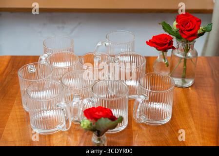 Ein elegantes Arrangement aus klaren Glasbechern auf einem Holztisch, begleitet von leuchtenden roten Rosen in kleinen Vasen, die eine raffinierte Tischszene schaffen. Stockfoto