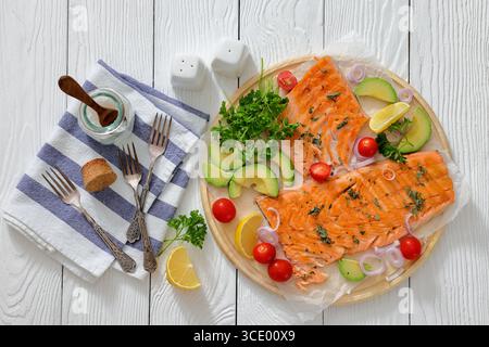 Gegrilltes Lachsfilet auf einem runden Holzbrett mit Zitrone, Avocado, Tomaten, Zwiebeln und Petersilie auf weißem Holztisch mit Gabeln und Serviette, Hori Stockfoto