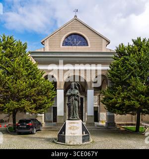 Izmir, Türkei - 6. April 2025: Außenansicht der St. John's Cathedral. Mit der Statue des Heiligen Johannes des Evangelisten, dem kopfsteingepflasterten Innenhof und grünen Bäumen unter blauem Himmel Stockfoto