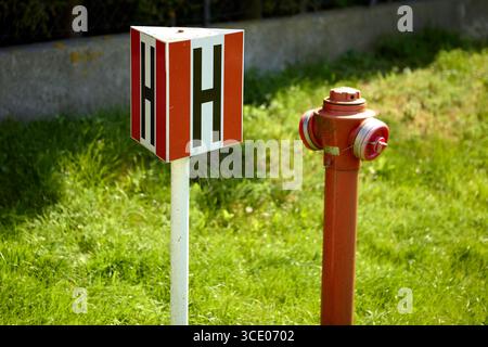 Roter Hydrant neben einem weißen Markierungspfosten mit roten Seiten und schwarzem H-Symbol, der auf grünem Gras in der Nähe einer Betonaufkantung installiert ist und die Wasserquelle anzeigt Stockfoto