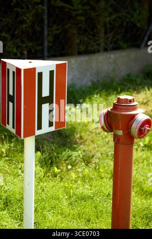 Roter Hydrant neben einem weißen Markierungspfosten mit roten Seiten und schwarzem H-Symbol, der auf grünem Gras in der Nähe einer Betonaufkantung installiert ist und die Wasserquelle anzeigt Stockfoto