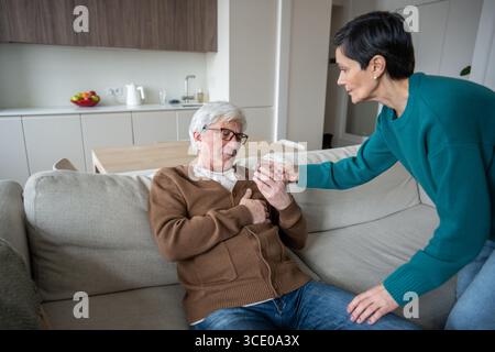 Die pflegende Ehefrau hilft älteren, leidenden Ehemann, der auf dem Sofa sitzt und ein Glas Wasser anbietet Stockfoto