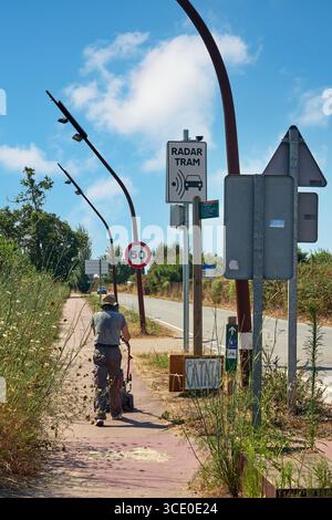 Viladecans. Barcelona - 14. August 2025: Ein Mann mit Strohhut schiebt einen Handwagen auf einen Bürgersteig. Die Szene, mit Verkehrsschildern und Vegetation, Illustration Stockfoto