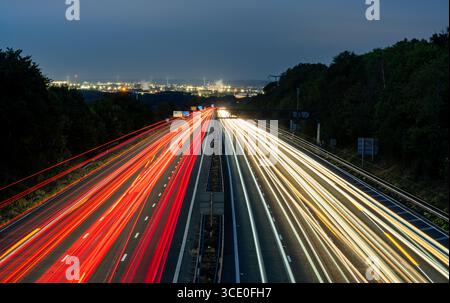 Langbelichtungsfoto mit lebendigen Lichtspuren auf einer Autobahn bei Dämmerung, mit der beleuchteten Stadt- und Industrielandschaft im Hintergrund Stockfoto