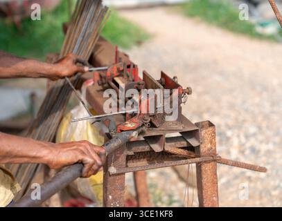 Nahaufnahme eines Arbeiters, der Stahlstäbe manuell mit einer rostigen Bewehrungsbiegemaschine auf einer Baustelle biegt. Stockfoto