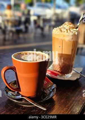 Ein frisch gebrühter Cappuccino in einer Tasse mit Untertasse, Teelöffel und Zucker und sitzt neben einem hohen Glas Eiskaffee mit Schlagsahne. Stockfoto