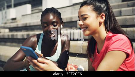 Sport, Ohrhörer und Freundinnen im Freien mit Telefon zum Musikhören, Netzwerken oder sozialen Medien. Lachen, Fitness und weibliche Athleten Stockfoto
