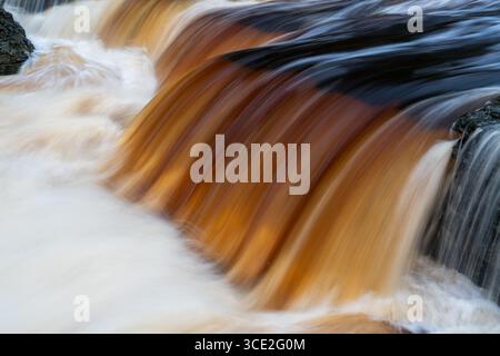 Nahaufnahme von braunem Flusswasser, gefärbt von Torfablagerungen, die in glattem Fließverhalten über Felsen kaskadieren Stockfoto
