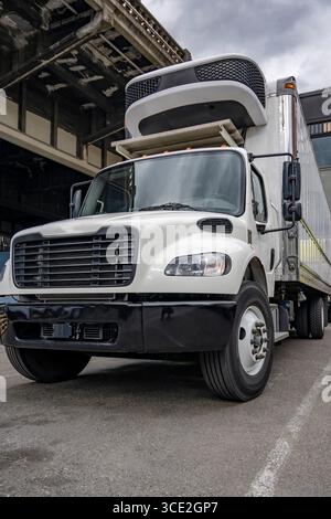 Industrieller mittelschwerer kompakter weißer Day Cab Sattelzugmaschine mit großem Lkw und Kühlkoffer Anhänger, der auf dem Industrieparkplatz steht und wartet Stockfoto
