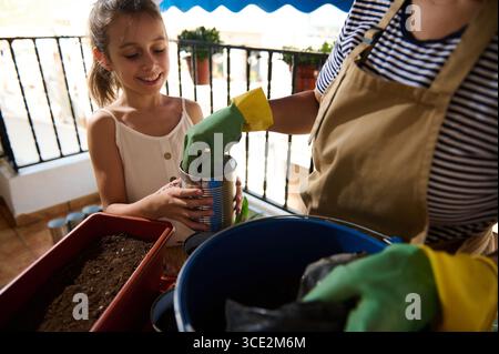 Eine Frau und ihre Tochter teilen sich einen Moment, indem sie Samen in Töpfen auf ihrem Balkon Pflanzen. Warmes Sonnenlicht und Teamwork fesseln die Beziehung und Freude der Familie Stockfoto