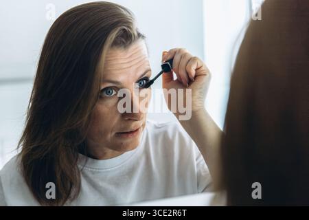 Frau mittleren Alters, die Augen mit einem Pinsel vor einem Badezimmerspiegel schminkt. Schönheitsroutine und Körperpflege im häuslichen Alltag. Hohe Qual Stockfoto