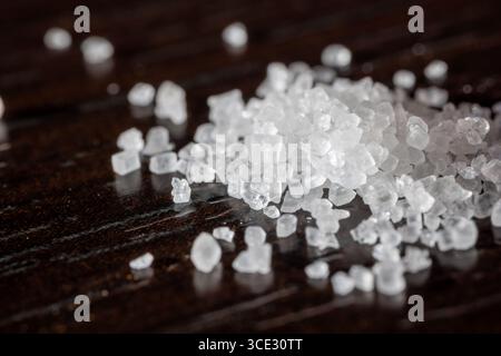 Makroaufnahme von groben Meersalzkristallen, die auf einer dunklen Holzoberfläche verstreut sind. Die Nahaufnahme zeigt die durchscheinende, kantige Textur der Salzkörner, e Stockfoto