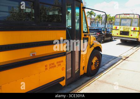 Nahaufnahme eines hellgelben Schulbusses, der entlang einer Stadtstraße unter sonnigem Himmel geparkt ist, mit einem weiteren Schulbus im Hintergrund. Stockfoto
