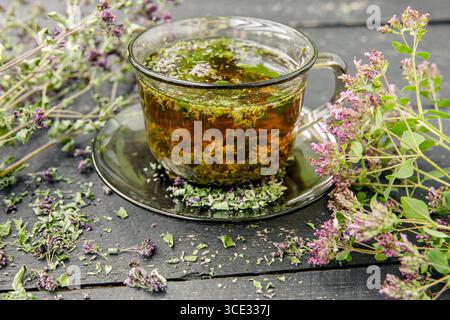 Hausgemachter Oregano, Origanum vulgare Kräutertee in einer Teetasse. Dampfendes Heißgetränk mit trockenen und frischen Oregano-Blüten rund um die Uhr, schwarzes Holz. Stockfoto