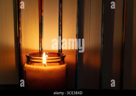Eine zündete Kerze in einem Glasgefäß vor Büchern. Die warme Flamme wirft ein gemütliches Leuchten auf den Buchrücken Stockfoto