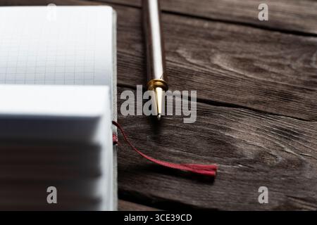Ein Notizbuch aus Raster liegt offen auf einem rustikalen Holztisch, begleitet von einem eleganten schwarzen Stift und einem roten Lesezeichen, das die warmen Texturen von W hervorhebt Stockfoto