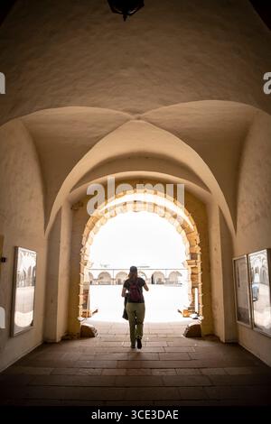 Ein Rückblick auf eine Person, die durch einen historischen, gewölbten Steinbogen zu einem sonnendurchfluteten Kopfsteinpflasterhof in einem klassischen Gebäude geht. Stockfoto