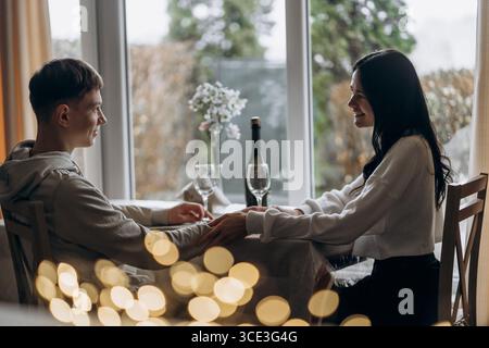 Romantische Winterferien, die Hände an einem gemütlichen Tisch mit Weingläsern und festlichen Bokeh-Lichtern halten, romantische Winterferien im Innenbereich mit warmem Wetter Stockfoto