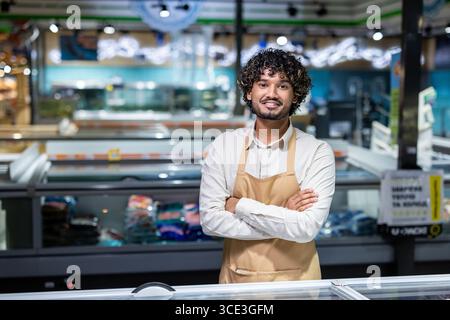 Ein lächelnder Mitarbeiter in Uniform, der selbstbewusst im Tiefkühlbereich eines Supermarktes steht. Kundenservice, Professionalität und eine saubere Einkaufsumgebung Stockfoto