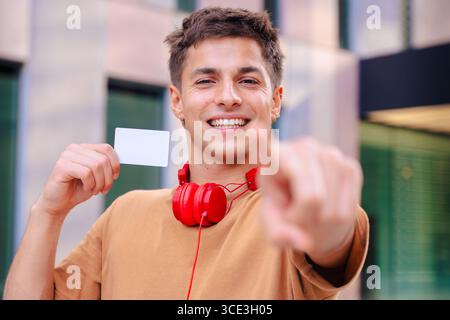 Ein lächelnder junger Mann, der auf die Kamera zeigt und draußen eine leere Visitenkarte hält Stockfoto