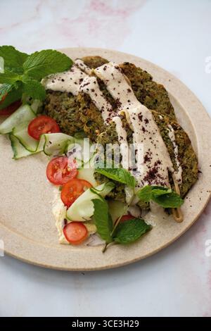 Teller mit knusprigen Falafelpasteten, serviert mit dünnen Gurkenscheiben, cremiger Tahini-Sauce, frischen Minzblättern und saftigen Kirschtomaten. Stockfoto