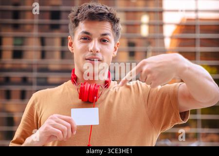 Ein lächelnder junger Mann, der auf die Kamera zeigt und draußen eine leere Visitenkarte hält Stockfoto