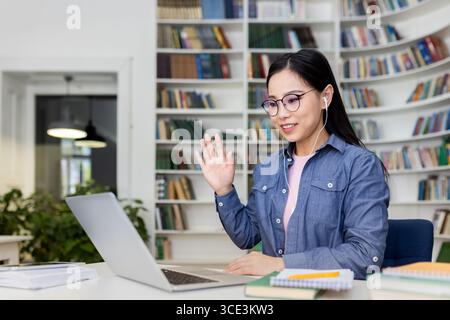 Eine junge Frau nimmt an einem virtuellen Meeting mit einem Laptop Teil, der in einer Bibliothek sitzt. Sie trägt Kopfhörer, winkt und wirkt in einer lebendigen digitalen Interaktion. Stockfoto