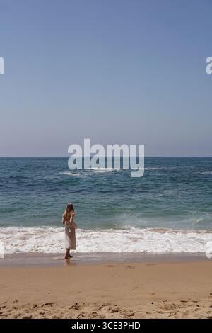 Eine Frau steht fröhlich an der Küste und genießt die beruhigenden Wellen an einem schönen sonnigen Strandtag Stockfoto
