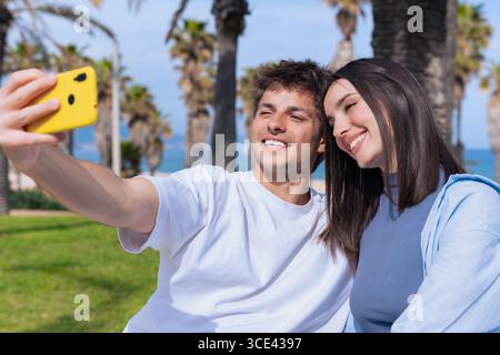 Verspieltes junges Paar, das Selfie mit gelbem Telefon am palmengesäumten Strand macht Stockfoto