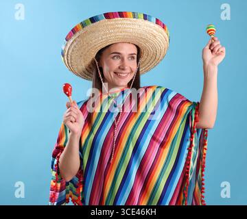 Glückliche Frau in Sombrero-Hut und farbenfrohem Poncho, der Maracas auf hellblauem Hintergrund spielt Stockfoto