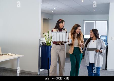 Verschiedene weibliche Kollegen, die durch den Büroflur laufen und dabei Laptop-Tablets und Kaffeetassen tragen Stockfoto