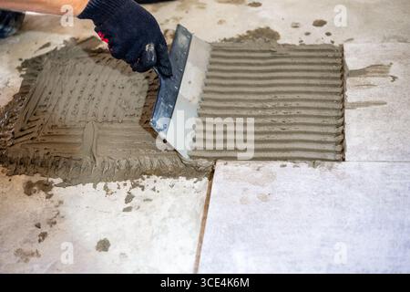 Ein Bauarbeiter trägt Klebermörtel mit einer gekerbten Kelle auf, bevor er Keramikfliesen auf einen Boden legt. Stockfoto