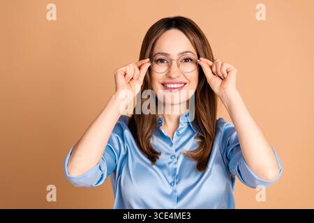 Selbstbewusste Geschäftsfrau mit Brille in blauer formeller Bluse, die vor beigefarbenem Hintergrund sitzt und Eleganz darstellt Stockfoto