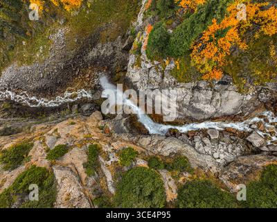 Eine kreative Perspektive von oben nach unten auf einen kleinen Wasserfall und Bach, der sich seinen Weg durch eine felsige Schlucht macht, im Kontrast zu leuchtendem orangefarbenem Herbstlaub Stockfoto