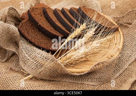 Nahaufnahme von geschnittenem Roggenbrot mit Koriandersamen auf einer Korbplatte mit Weizenohren über Sackleinen Stoff rustikales Lebensmittelkonzept Bäckerei-Landwirtschaft Stockfoto