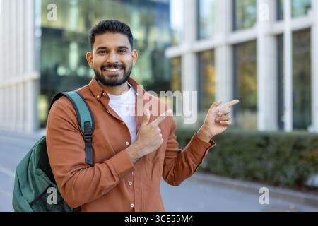 Junger Mann mit Rucksack, lächelt und zeigt selbstbewusst, gefangen in einer städtischen Umgebung. Stockfoto