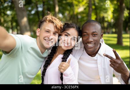 Multirassische Studenten befreunden sich, Selfie zu machen, V-Zeichen zu gestikulieren und Daumen hochzuzeigen, im Freien auf dem Campus zu posieren Stockfoto