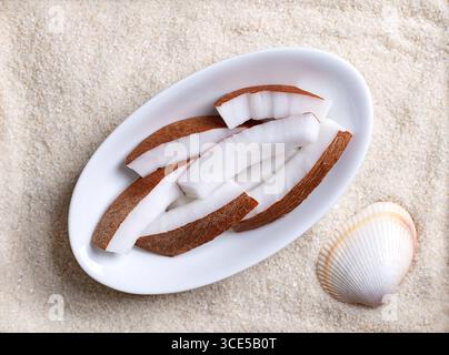 Frisches Kokosfleisch, in ovaler weißer Schüssel, auf Strandsand mit Schale. Deckendes weißes Fleisch einer frischen und jungen Kokosnuss, in Streifen geschnitten und verzehrfertig. Stockfoto