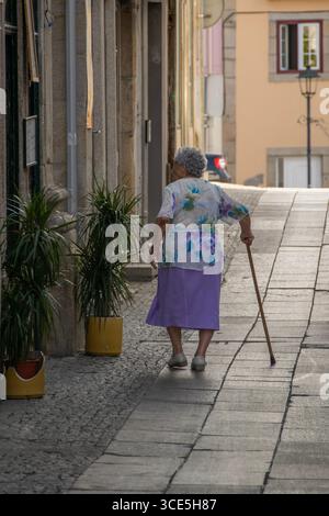 Chaves, Portugal: 13. August 2023: Ältere Frau mit einem Stock, die in einer engen Straße im historischen Zentrum von chaves, portugal, spaziert Stockfoto