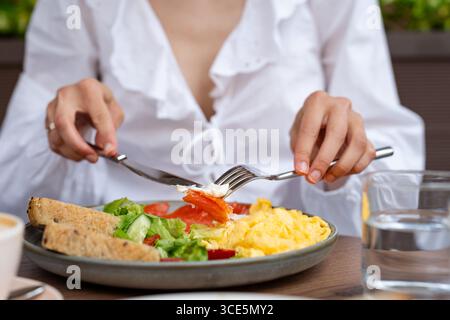 Nahaufnahme einer jungen Frau, die an einem Tisch sitzt und geräucherten Lachs, Rührei, Salat und Brotscheiben mit einem Glas Wasser isst Stockfoto