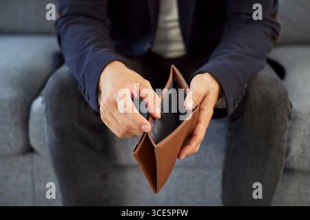 Ein armer Mann, der leere Geldbeutel hält und öffnet, ohne Geld zu Hause auf dem Sofa zu sitzen. Stockfoto