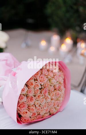Luxuriöser großer Strauß rosafarbener Rosen, gewickelt in weichem rosa Papier, liegt auf einem Tisch, im Gartenhintergrund mit verschwommenen Glühkerzen. Romantisch und Stockfoto