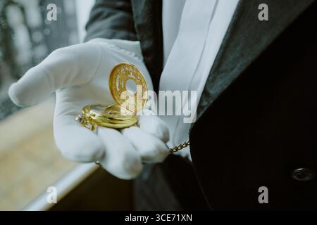 Kaukasischer Mann mittleren Alters, der einen formellen Anzug trägt, der eine goldene Taschenuhr in Handschuhen hält und drinnen in der Nähe des Fensters steht und komplizierte Uhrendetails zeigt Stockfoto