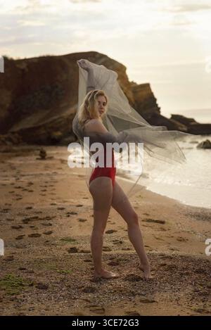 Helle und junge Frau unbeschwert genießt die Sonne an einem schönen Tag am Strand, tanzt fröhlich in einem roten Badeanzug mit fließendem Stoff. Die blonde c Stockfoto