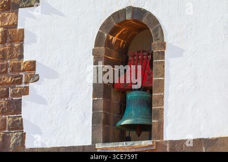 Architektonische Details mit Glocke, die von einem Steinbogen eingerahmt wird, vor dem Hintergrund einer weiß getünchten Wand. Bronzeglocke mit grüner Patina hängt in einem Stein Stockfoto