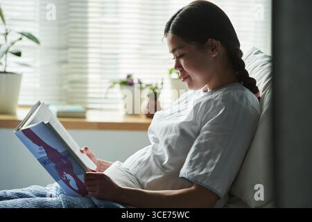 Junge Frau, die sich drinnen entspannt, während sie im Morgenlicht das Schwangerschaftsbuch liest Stockfoto