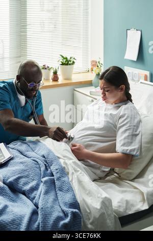 Schwarzer männlicher Arzt mittleren Alters, der schwangere Kaukasierin bei Geburt untersucht Stockfoto