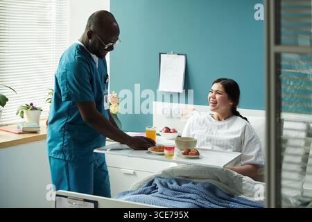 Schwarze männliche Krankenschwester serviert Frühstück für kaukasische Frau im Entbindungskrankenhaus Stockfoto